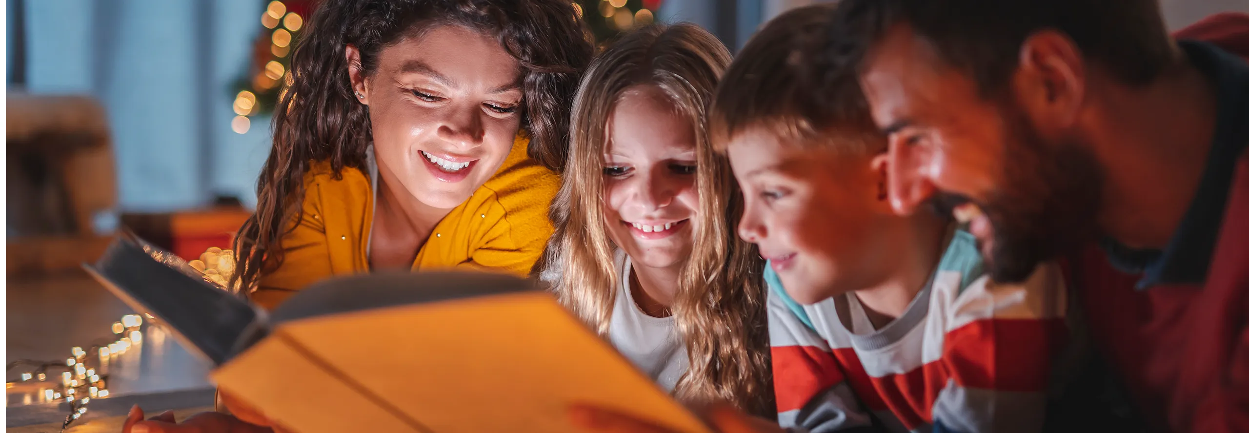 Família (Mãe, Pai e dois filhos) a ler no chão com árvore de natal em fundo